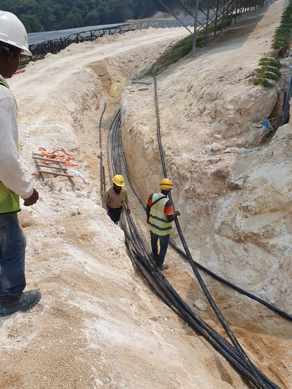 Workers guiding multiple cables into an open trench on the hillside, with solar panel structures visible above.